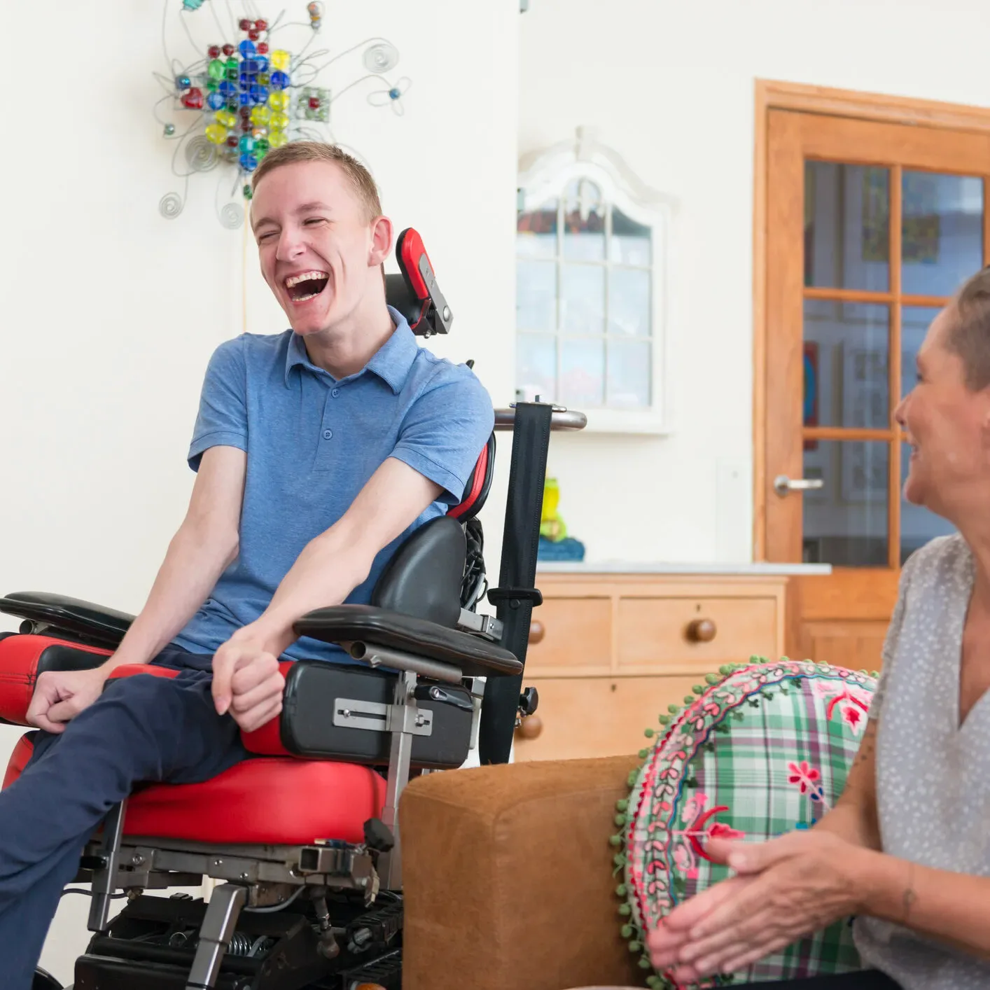 Young man in a power wheelchair laughing with his caregiver at home
