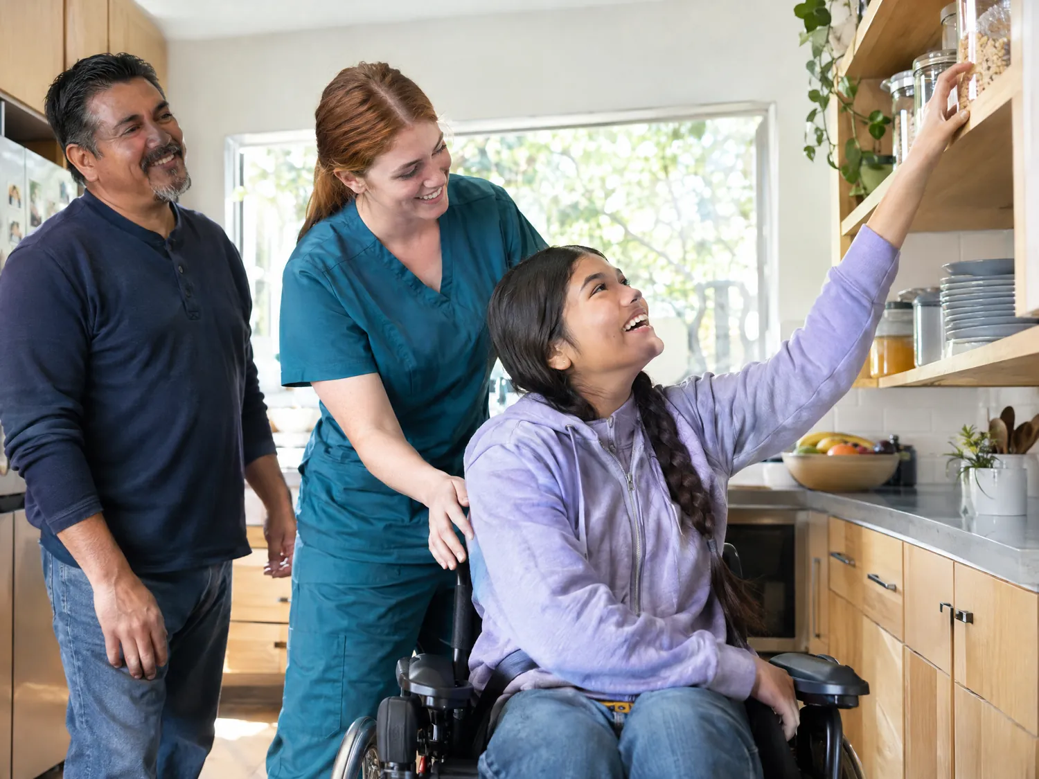 Caregiver helping a laughing teen in a wheelchair reach a kitchen shelf